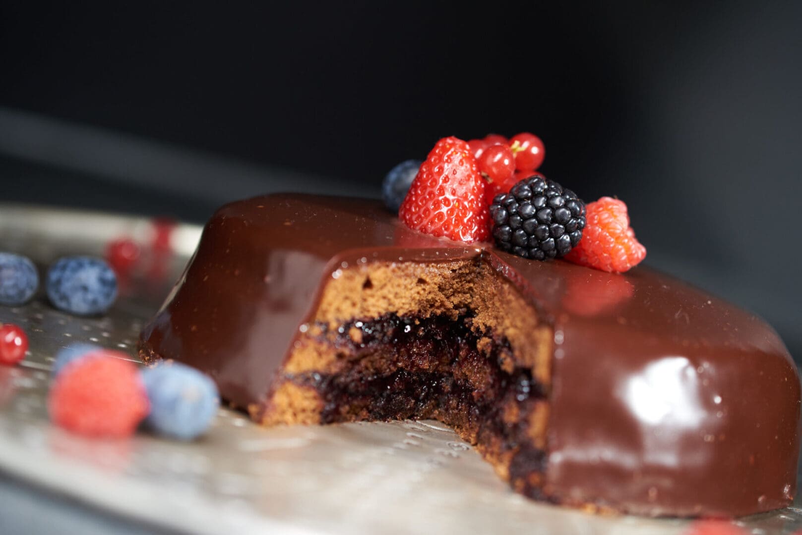 A chocolate-glazed cake with a slice removed, showing a dark berry filling. The cake is topped with fresh strawberries, blackberries, and redcurrants. Additional berries surround the cake on the gray surface.