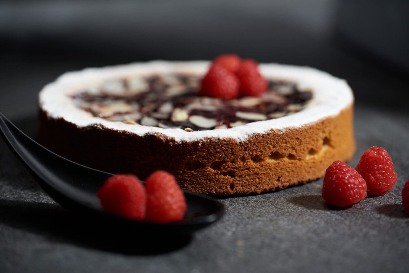A round cake with a golden-brown crust and powdered sugar on top, garnished with fresh raspberries. Additional raspberries are placed on a black spoon and nearby on a gray surface.