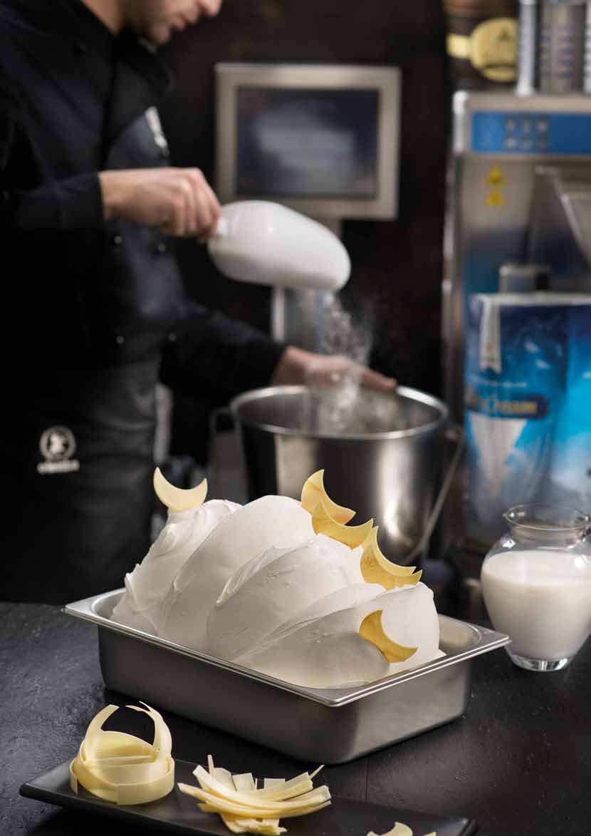 A person in a black uniform is pouring milk into a large metal mixing bowl, crafting the perfect ice cream base using Elenka Ice Cream Base Ingredients Novacrem 50. In the foreground, a container of smooth, white gelato garnished with decorative white chocolate curls is placed on a dark surface. A jug of milk and a tray with more white chocolate curls are nearby.