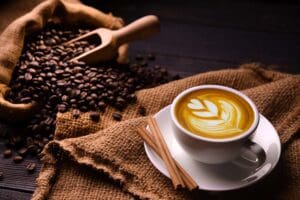 A cup of coffee with latte art sits on a white saucer beside cinnamon sticks, evoking the warmth of a cozy grocery aisle. Coffee beans spill from a wooden scoop onto burlap cloth, creating a rustic charm, reminiscent of fresh finds during food shopping. The scene is warmly lit, highlighting rich tones.
