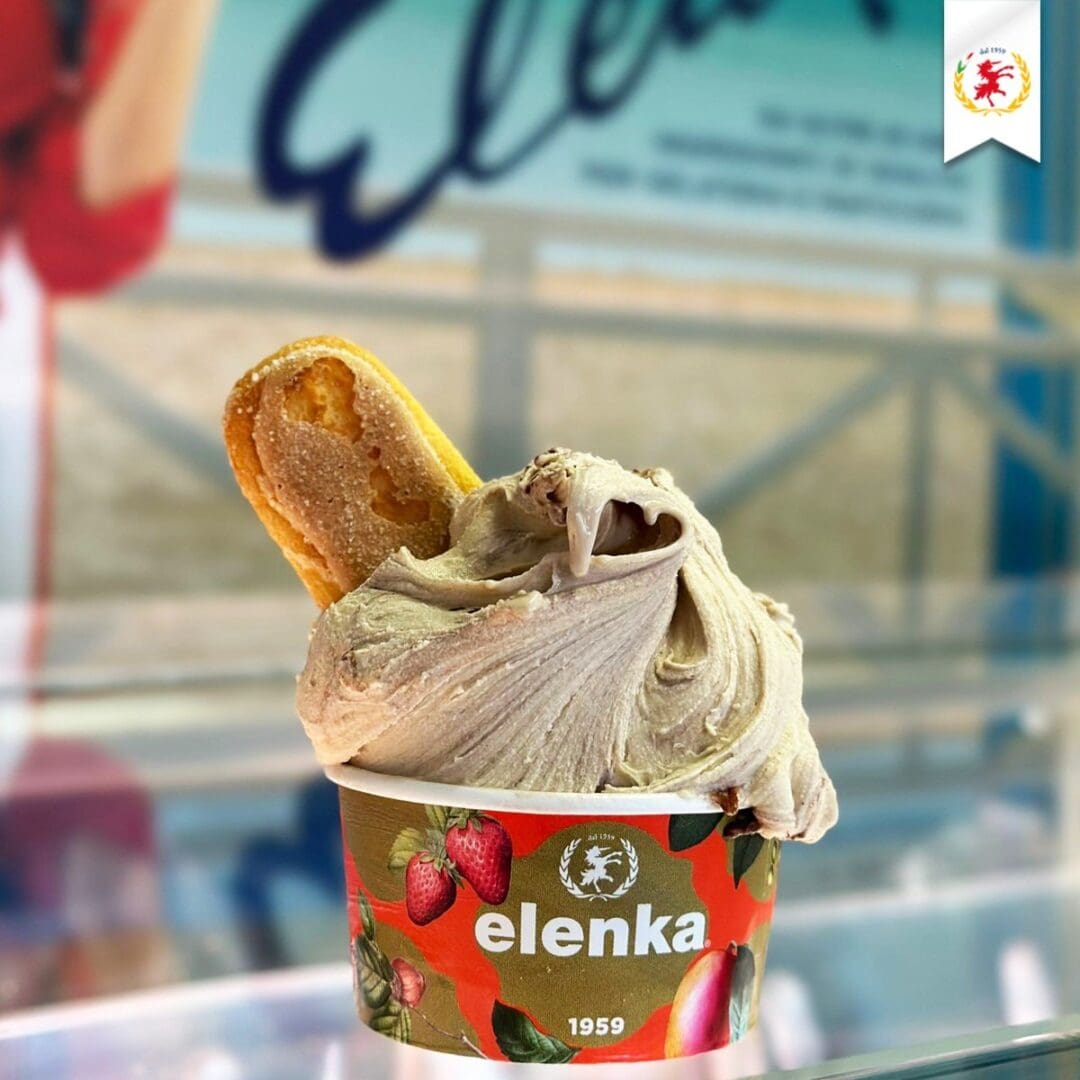 A cup of gelato topped with a biscuit, set against a blurred background. The cup features "elenka" branding with strawberry and branding graphics. The gelato is creamy with a swirl, and the biscuit is partially inserted on one side.