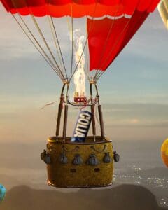 Golia Hard Balsamic Candy and A hot air balloon floats in the sky with a basket holding a large cola can. The balloon's vibrant red canopy contrasts with the soft, scenic backdrop of mountains and a city below. Bright flames are visible above the cola can.