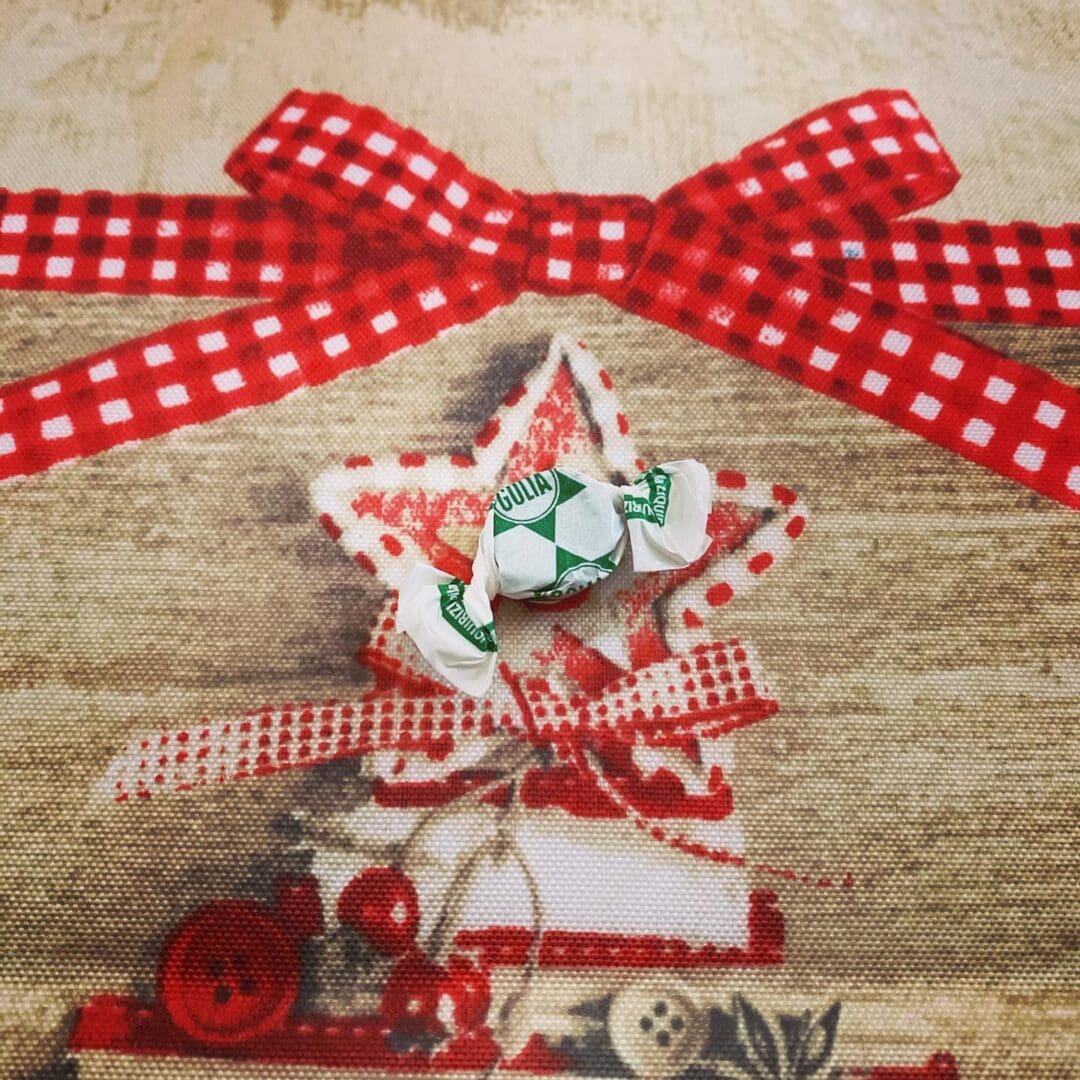 A wrapped Golia Licorice Gummy Candy with green and white packaging rests on festive fabric, backed by a star decorated with red and white checkered ribbons and red embellishments, including a button.