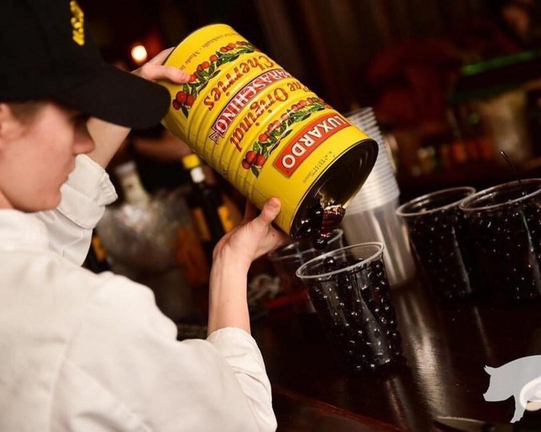 A person in a black cap pours black olives from a large yellow can into a plastic cup on a dark wooden surface. Nearby, several filled and stacked white cups sit alongside Luxardo The Original Maraschino Cherries (4x5.6Kg case), adding elegance to the scene.