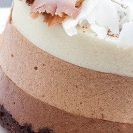 Close-up of a triple-layered chocolate mousse cake. The dessert features layers of dark, milk, and white chocolate mousse, topped with chocolate shavings in brown and white. The base is a dark chocolate crust.