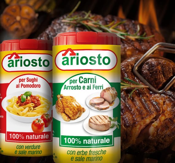 Two containers of Ariosto seasoning mix are displayed in front of a grilling steak. One container is for tomato sauces, and the other is for roasted meats. Both are labeled as "100% naturale" with various foods depicted on the packaging.