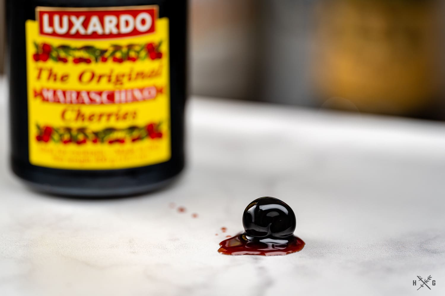 A single maraschino cherry sits on a white marble surface, with its syrup trailing behind. The blurred background features a Luxardo jar labeled "The Original Maraschino Cherries.