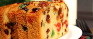 Close-up of a sliced fruitcake on a white plate. The cake is filled with colorful candied fruits, nuts, and raisins, showcasing a moist, dense texture. Forks are visible in the background.