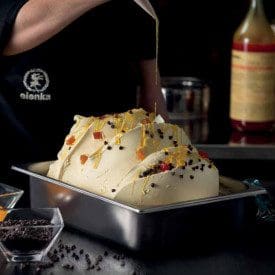 In a professional kitchen, a chef drizzles syrup over a large gelato container topped with fruit and chocolate chips, enhanced by Elenka (303) Zuppa Inglese Extract (6x2.75Kg case), creating an irresistible masterpiece.