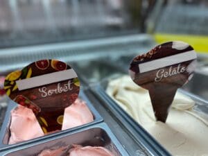 Close-up of an ice cream display featuring two flavors. The left tub contains pink sorbet with a sign reading "Sorbet" and fruit imagery. The right tub holds a pale gelato labeled "Gelato" with a wooden-themed sign.