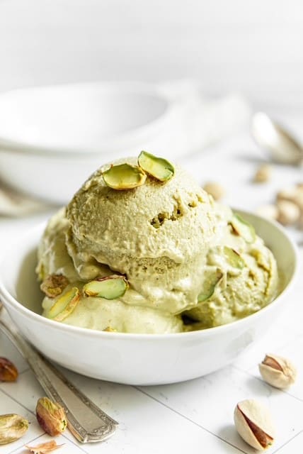 A bowl of creamy Elenka Pistachio P ice cream (4x2.5Kg), topped with sliced pistachios, sits on a white surface amidst scattered nuts and silver spoons, epitomizing indulgence.