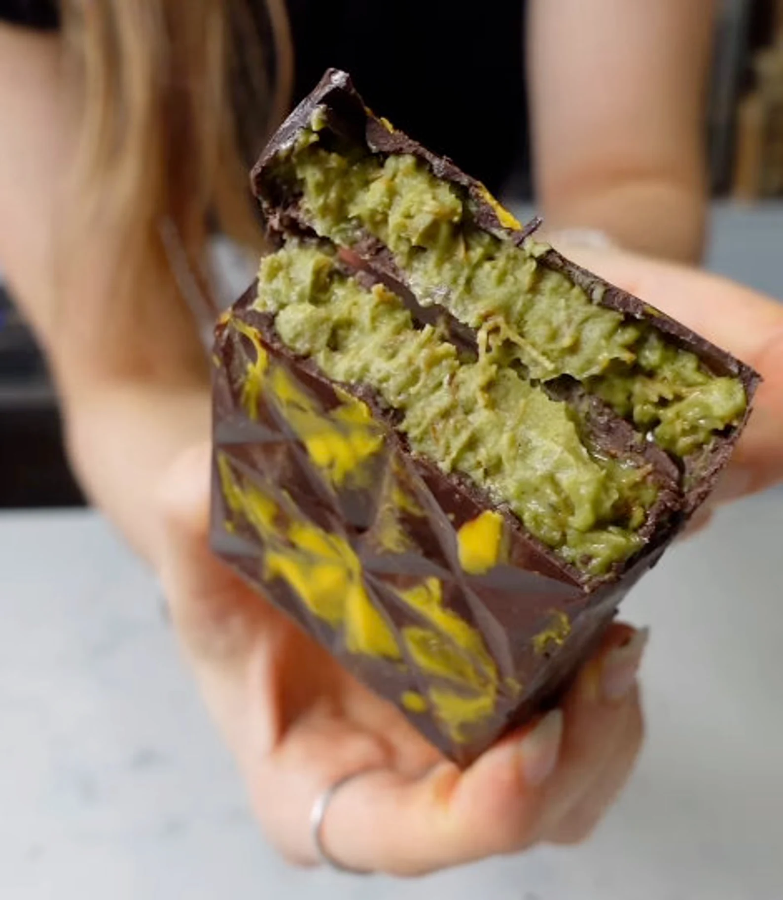 Close-up of a person holding an Elenka Pistachio Premium Nuts Variegato (Dubai Chocolate Bar) featuring triangular patterns on its exterior. The inside reveals a green pistachio filling, contrasting with the brown chocolate. The person's hand and part of their hair are visible.