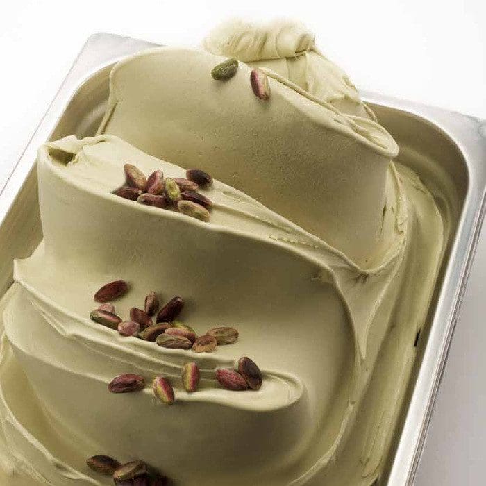 A tray of pistachio gelato, crafted with Elenka (775) Pistachio Pure Paste for Ice-Cream, features a swirl of whole pistachios atop its creamy surface. The pale green gelato contrasts beautifully with the brown nuts for an elegantly appealing dessert presentation.