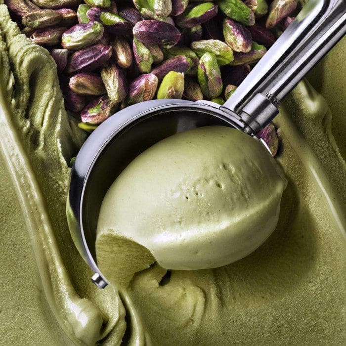 A metal scoop lifts a smooth scoop of creamy green pistachio ice cream made with Elenka (775) Pistachio Pure Paste. Whole pistachios in the background emphasize the flavor, perfectly complementing the ice cream.