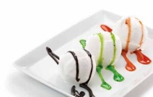 Three scoops of vanilla ice cream on a white rectangular plate, Elenka Toppings drizzled with chocolate, green, and red syrups, creating a colorful presentation against a plain white background.