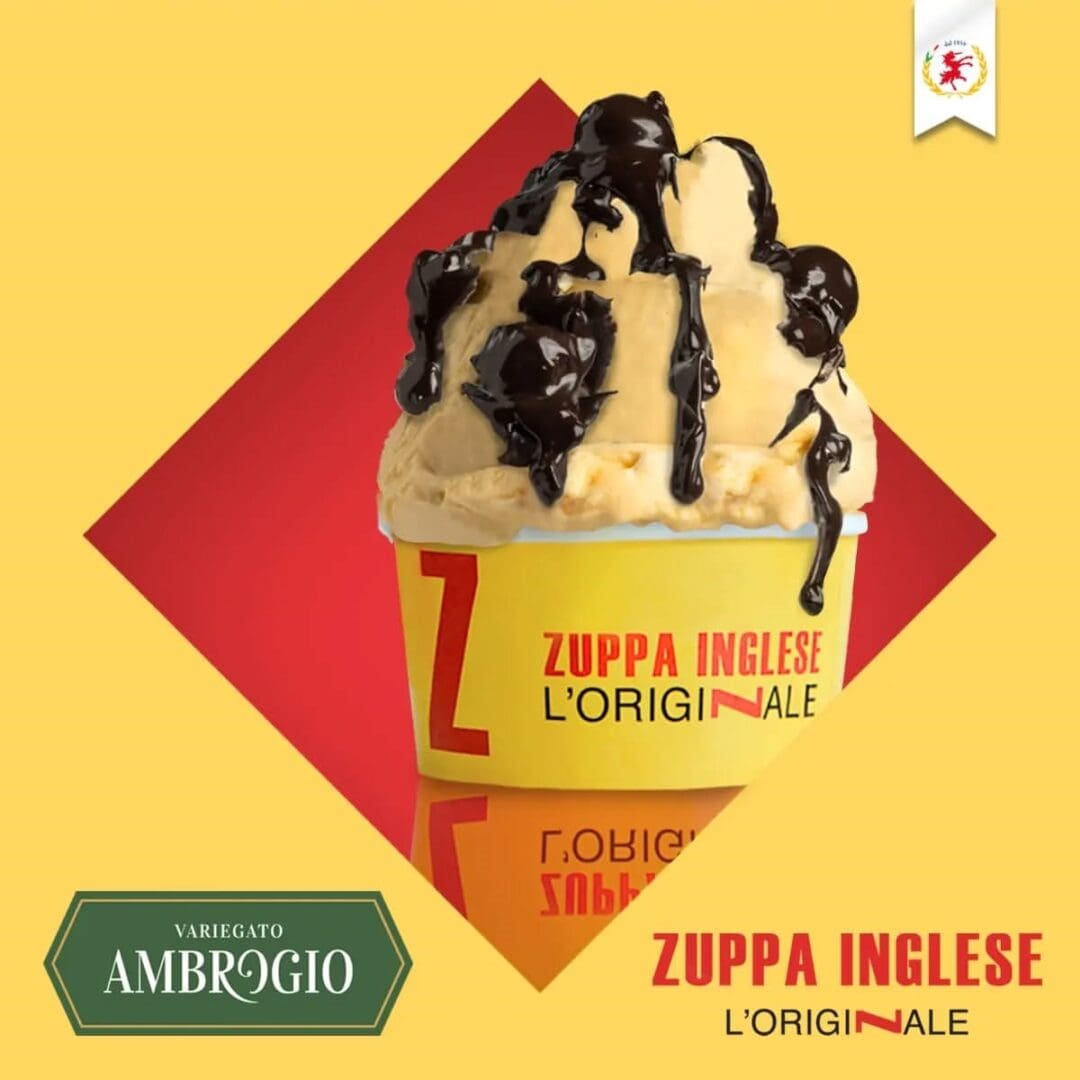 A cup of gelato featuring Elenka Zuppa Inglese Extract, drizzled with chocolate syrup, in a yellow cup with red text. It showcases "Variegato Ambrogio" and "Zuppa Inglese L'Originale" logos on a yellow diamond background.