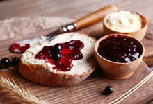 A wooden board features a slice of bread with butter and Luxardo Raspberry Jams and Spreads (4x5.4Li case) surrounded by small wooden bowls filled with more butter and jam, scattered berries, and a stalk of wheat for a rustic touch.
