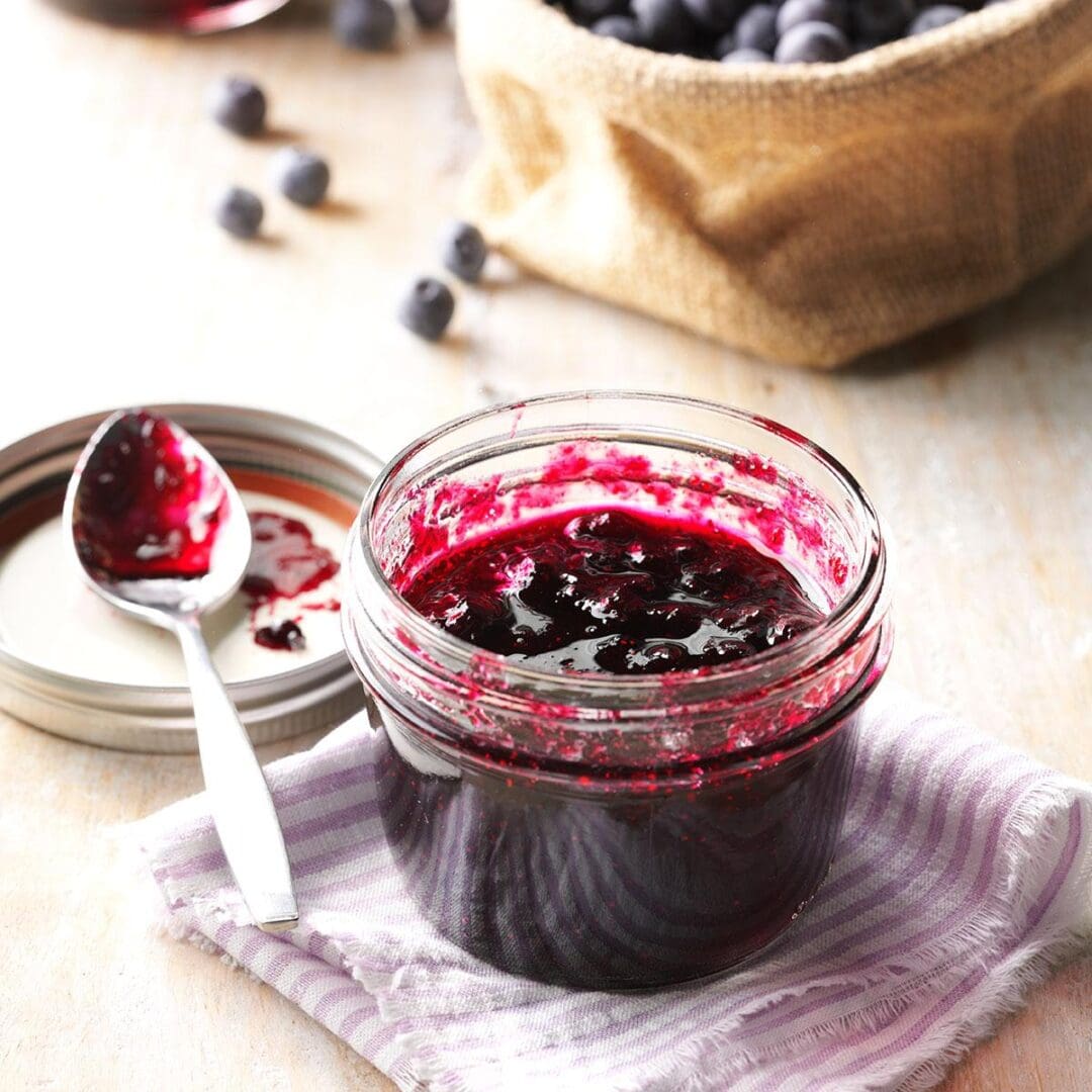 A jar of Luxardo Premium Blueberry Jams and Spreads 'Mirtillo' is on a cloth, with an open lid and jam-covered spoon nearby. Blueberries are scattered in the background alongside a burlap sack full of berries.