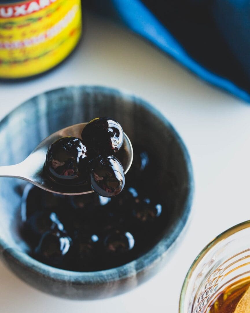 A spoon holds shiny black tapioca pearls above a bowl of more pearls, similar to Luxardo Burgundy Red Cherries. In the background, a blurred bottle with yellow and red labeling is next to a glass of rich brown liquid.