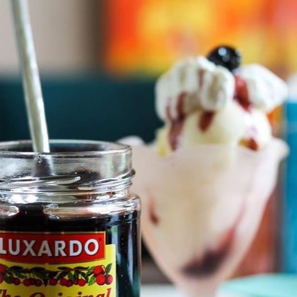 A jar labeled "Luxardo Gourmet Maraschino Cherries" is close to a glass dish sundae topped with whipped cream, a cherry, and syrup. The softly blurred warm colors in the background enhance the decadent scene.