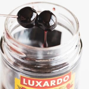 A close-up shows a jar labeled "Luxardo Gourmet Maraschino Cherries," with two cherries on a metal skewer being lifted from the syrup. The label showcases an elegant cherry design and text.