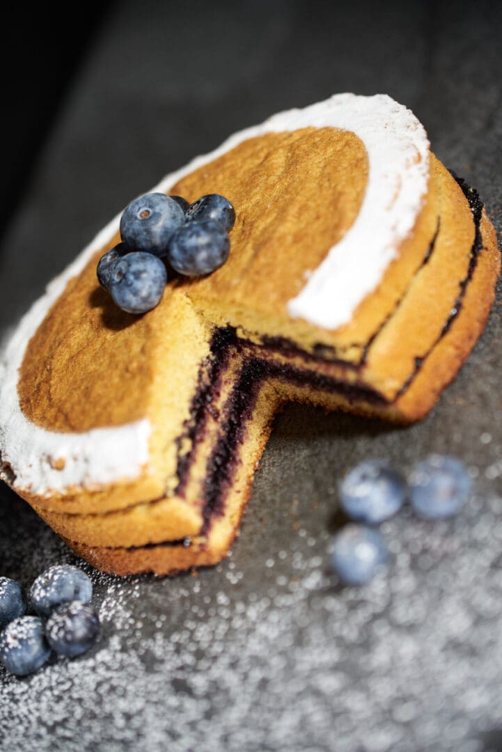 A round cake with a slice missing showcases rich Luxardo Premium Blueberry Jams and Spreads 'Mirtillo' filling. The top is lightly frosted, adorned with fresh blueberries, and surrounded by scattered berries on a dark surface.