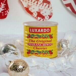 A case of Luxardo Burgundy Red Cherries enhances holiday cheer, surrounded by silver and gold glittery baubles and white string lights. The rich cherries add to the festive scene, with red and white stockings hanging in the background.