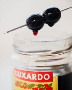 Two dark cherries on a cocktail pick hover above a jar labeled "Luxardo," with cherry illustrations visible. A drop of juice hangs from one cherry, suggesting a rich, syrupy texture. The background is softly blurred.