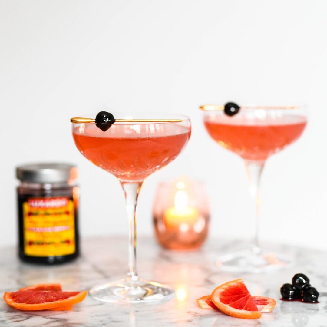 Two elegant cocktail glasses with pink drinks, garnished with Luxardo Gourmet Maraschino Cherries and gold rims, sit on a marble surface. A small candle glows nearby, alongside sliced blood oranges and a jar of Luxardo cherries.