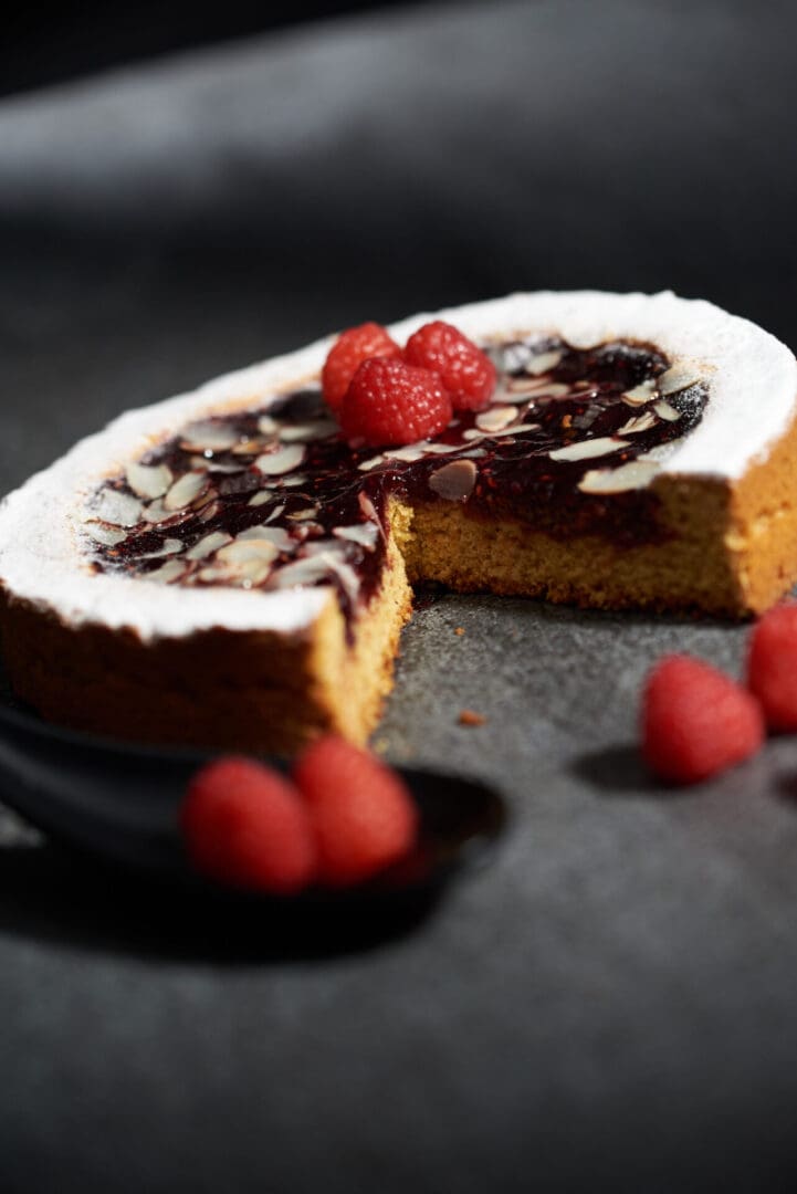 A round almond and raspberry tart with a slice missing reveals its moist interior. Topped with sliced almonds and fresh raspberries, it features a hint of Luxardo Raspberry Jams and Spreads (4x5.4Li), surrounded by extra berries on a dark surface next to a black spoon.