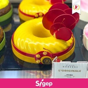 A decorative yellow cake with a glossy finish, featuring circular red accents and a red ribbon with flower designs. It is displayed on a tray with a sign reading "L'Originale" and the logo "Sigep" at the bottom.