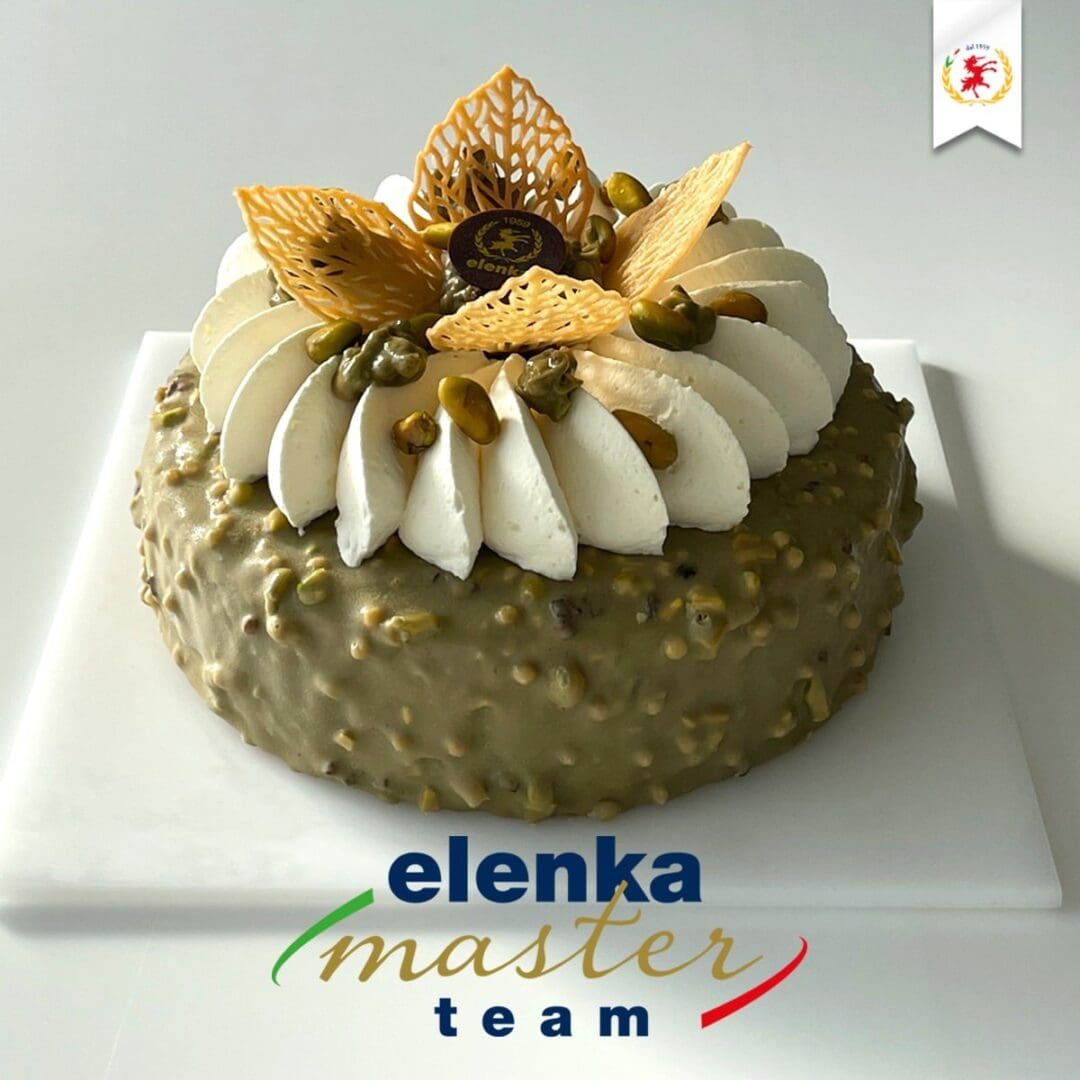 A cake on a white platter, covered in green icing with nuts and whispers of Luxardo Cherries. It’s topped with white cream swirls, decorative wafers, and nuts. There’s a small brown label on top. A logo reads "elenka master team" below the cake.