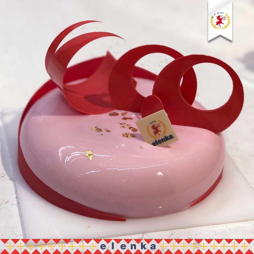 A heart-shaped cake with a glossy pink surface is adorned with red chocolate loops and gold flakes, reminiscent of Elenka Gelato's elegance. It rests on a white square board featuring a small "elenka" tag with a red emblem.