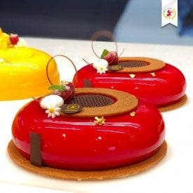 Two glossy red glazed cakes with circular chocolate decorations and small flowers on top, each adorned with a Luxardo Cherry. Resting on thin brown bases, these elegant creations are reminiscent of fine Elenka Gelato craftsmanship. A small emblem graces the top corner.