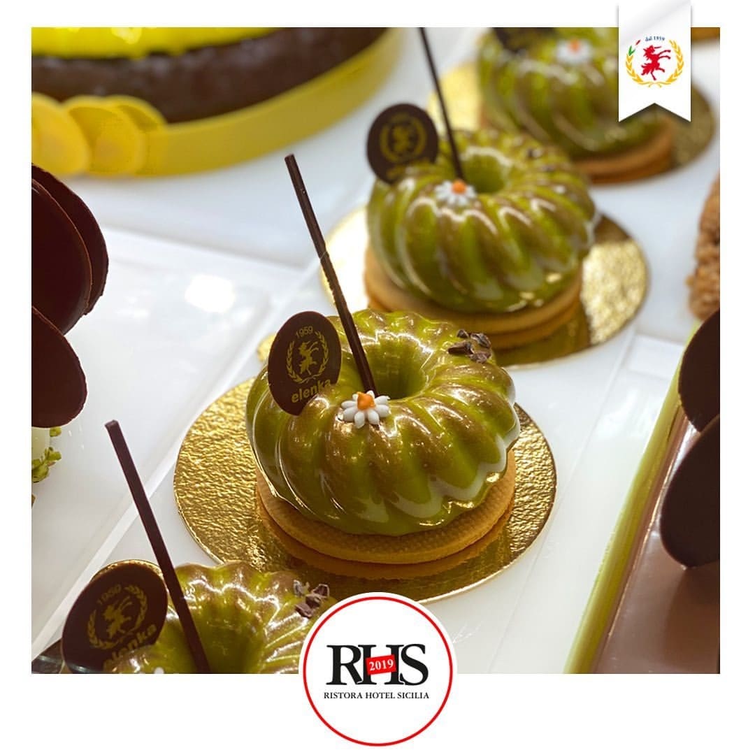 Elegant green glazed bundt-shaped pastries on golden bases, exuding sophistication, are topped with chocolate discs and delicate flowers. This delightful display perfectly complements the rich flavors of Elenka Gelato or the tangy sweetness of Luxardo Cherries in a bakery or dessert setting.