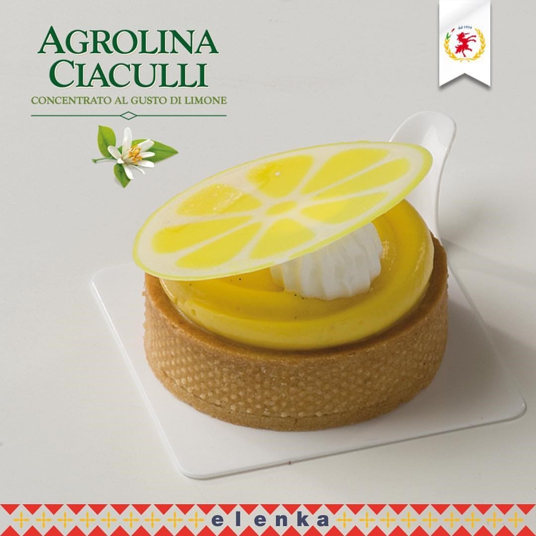 A lemon-flavored pastry with a glossy yellow filling, topped with a slice of decorative, translucent lemon. It sits on a white square base, adorned with "Agrolina Ciaculli" and “elenka” text, offering hints of Elenka Gelato's rich essence in every bite.
