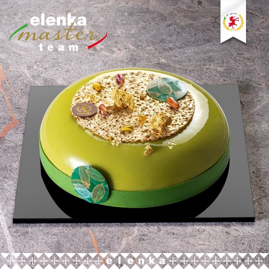 A round green mousse cake with a glossy finish, topped with beige decorative elements, pistachios, and circular designs. It's displayed on a black square board with logos and text "elenka master team" visible.