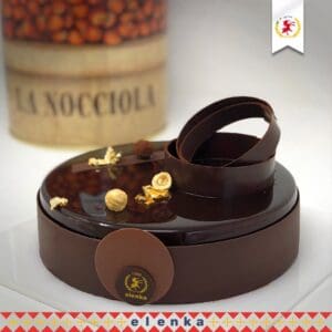A round, glossy chocolate cake is decorated with hazelnuts and gold leaf on top. Two circular chocolate pieces are elegantly placed on the cake. In the background is a container labeled "La Nocciola." The cake is on a white surface.