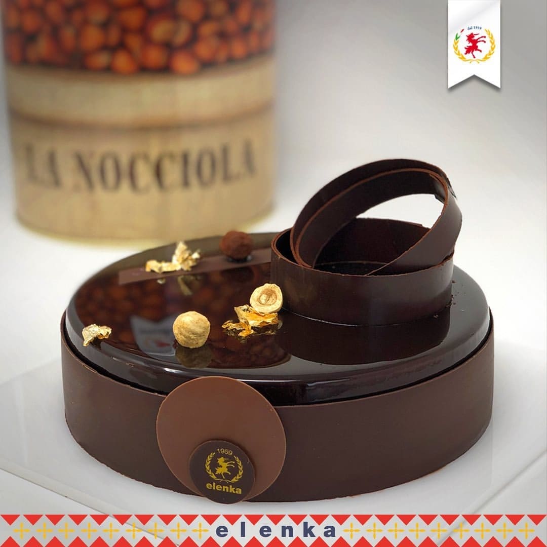 A glossy chocolate cake adorned with hazelnuts and gold leaf sits on a white plate, featuring sleek chocolate rings on top. In the background, a large container labeled "La Nocciola" hints at Elenka Gelato, while Luxardo Cherries add a pop of color among the row of vibrant triangles.