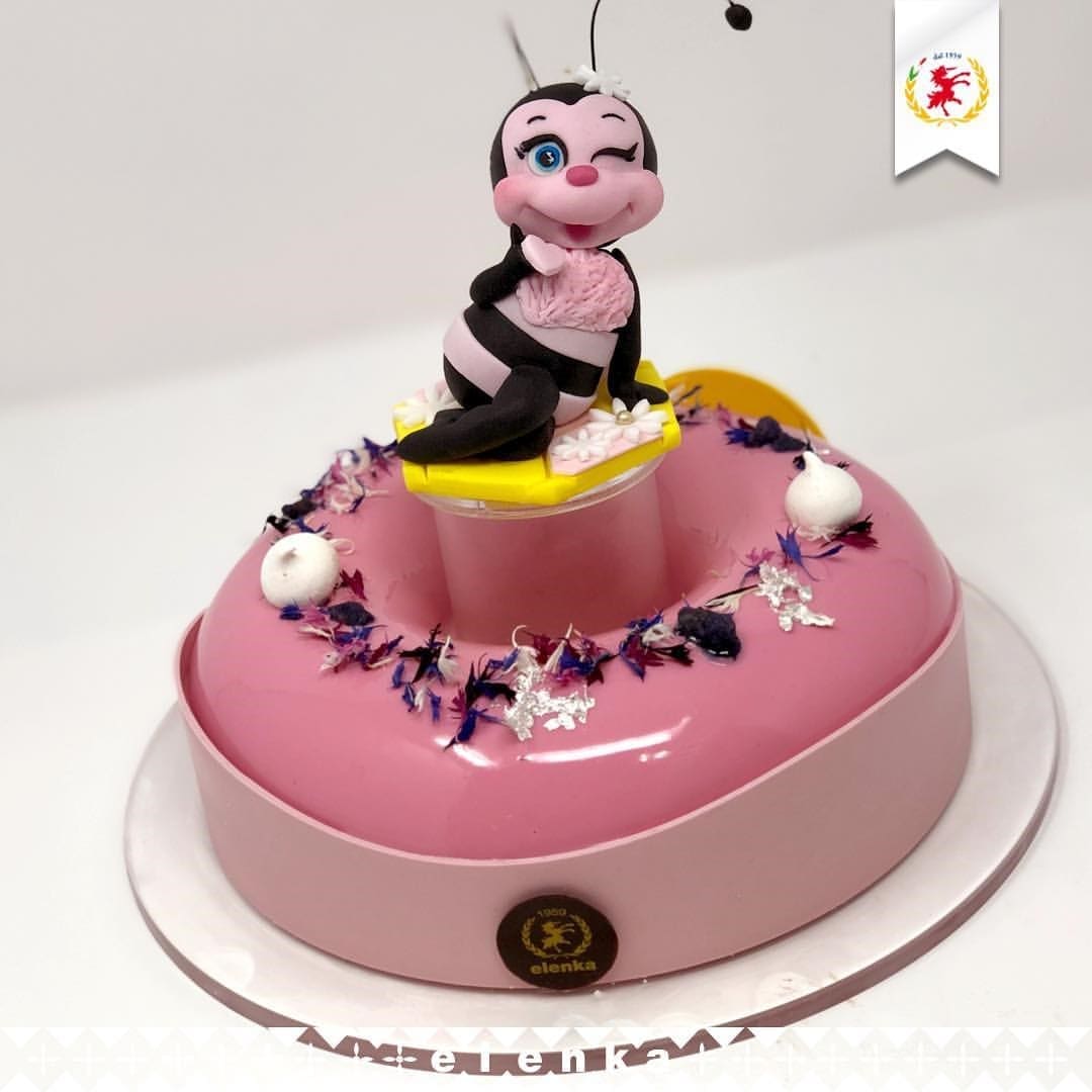 A pink heart-shaped cake with a glossy finish, adorned with edible flowers, Luxardo cherries, and two small spheres. A cheeky cartoon bee figure sits on top, winking mischievously. The cake features a small logo reading "Elenka Gelato.