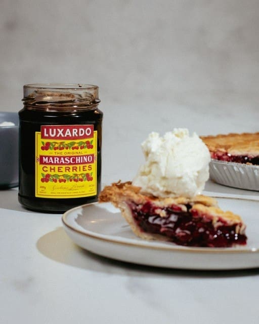 A slice of cherry pie with a scoop of Elenka Gelato vanilla ice cream graces a white plate. In the background, there's a jar of Luxardo maraschino cherries and another slice of pie waiting to be savored. The setting is on a light surface, capturing the essence of indulgence.