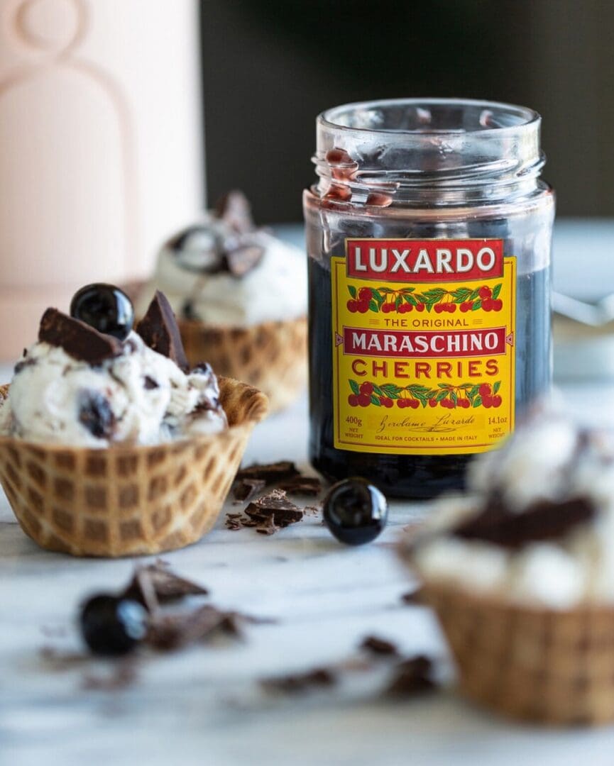 A jar of Luxardo maraschino cherries sits amidst waffle bowls brimming with rich Elenka Gelato, chocolate chunks, and cherries. Scattered pieces of chocolate adorn the cool marble surface.