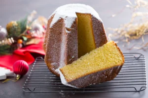 A tall, dome-shaped Pandoro cake dusted with powdered sugar is displayed on a cooling rack. A slice has been cut and placed to reveal the soft, yellow interior. Festive decorations and a red cloth are in the background.