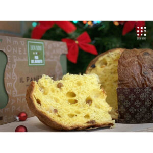 Close-up of sliced panettone with raisins, in front of a branded box. The background is festive with blurred holiday lights and a hint of a red ornament. The scene suggests a holiday setting.