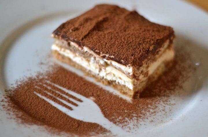 A slice of tiramisu rests on a white plate, dusted with Elenka (101) Cacao Amaro-Bitter Cacao powder (20x1Kg case), with a fork-shaped outline where the rich cocoa was artfully blocked.