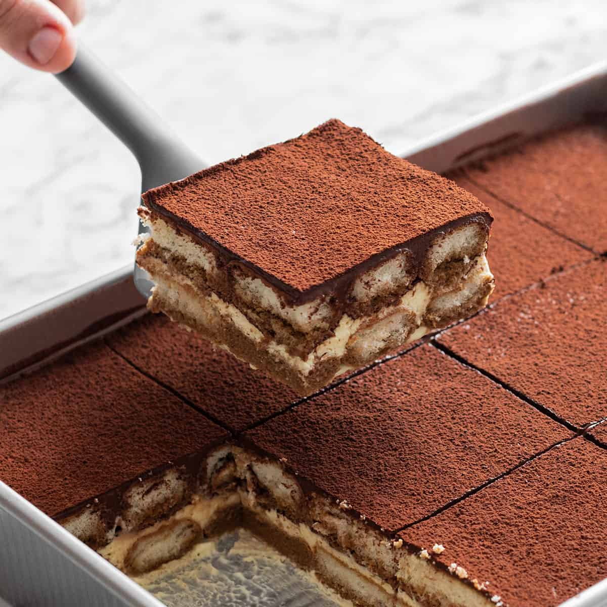 A hand lifts a square tiramisu from a pan, revealing coffee-soaked ladyfingers and creamy layers made with Elenka (443) Tiramisu Gelato Paste (2x6Kg case), topped with cocoa powder. More neatly cut pieces remain in the pan.