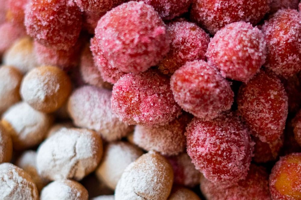 A close-up of red and orange fruits with sugar crystals, enhanced by Luxardo Alkermes Aroma 70% Alcohol (2x5Li case), sits beside round brown treats covered in powdered sugar, making a colorful, textured display.