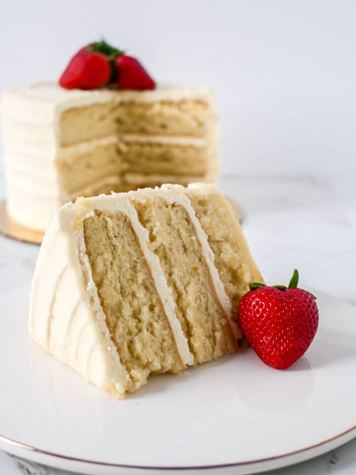 A slice of three-layer vanilla cake with white frosting sits beside a fresh strawberry, while the whole cake behind is topped with two strawberries and exudes an intense aroma like Luxardo Lacrime D'oro (Benevento) 70% Vol. Alcohol.