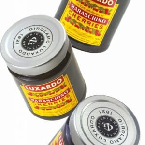 Three Luxardo Maraschino Cherries (400g jars) with red and yellow labels are arranged diagonally on a white background; two jars stand upright while one lies on its side.