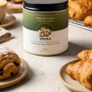 A jar from a 12x454g case of Elenka Pistachio Cream sits on a table surrounded by croissants, pastries, and a panettone in the background, creating a cozy breakfast scene.